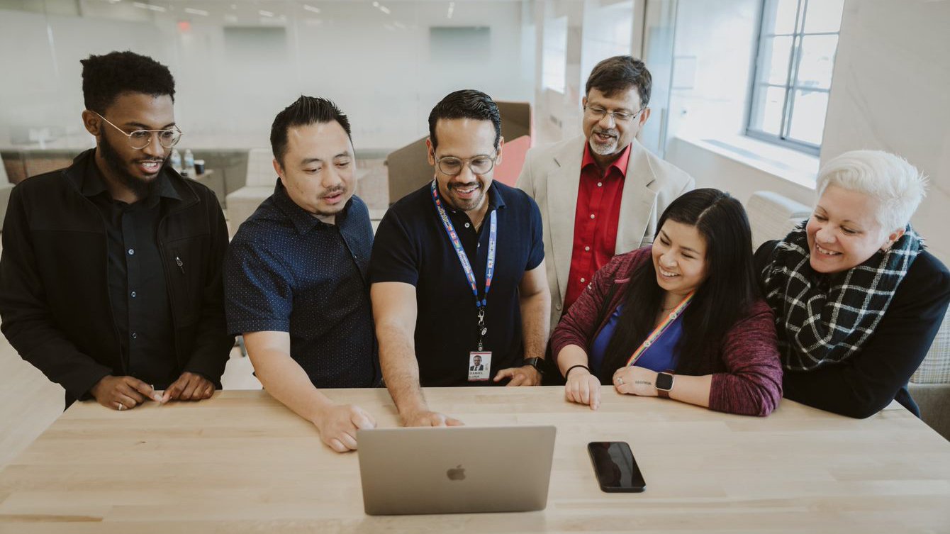 Diverse group of six people all smiling and looking at a laptop on a table.