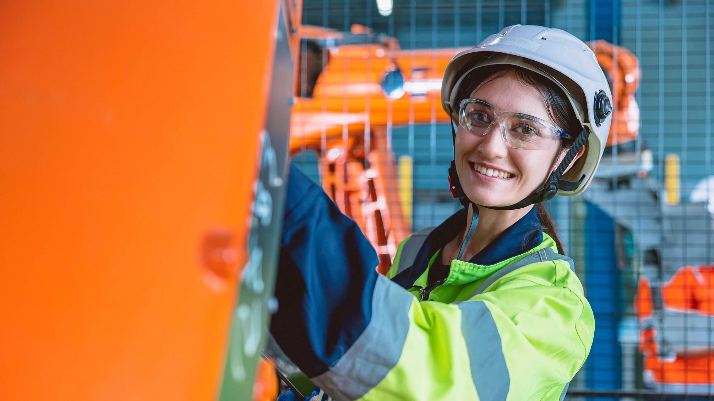 Young woman in factory wearing white hardhat, safety glasses and neon green coat is holding a laptop computer and checking electronic panel. Orange robotic arm is behind woman -- in manufacturing line.
