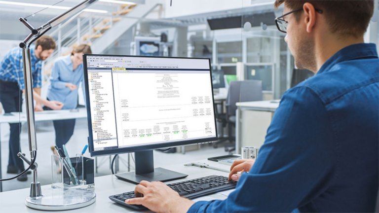 Un empleado con lentes viendo software en el monitor de su escritorio con dos empleados en segundo plano colaborando en otro proyecto