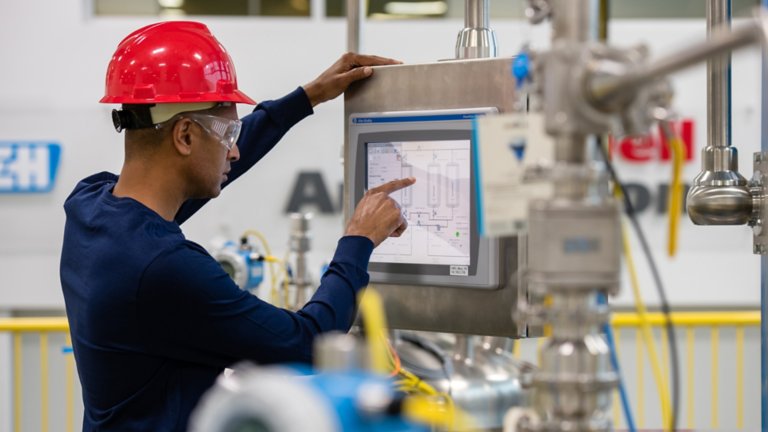一名戴红色安全帽的男性 Rockwell Automation 员工在工厂监视器的触摸屏上将信息输入 FactoryTalk View 软件应用程序