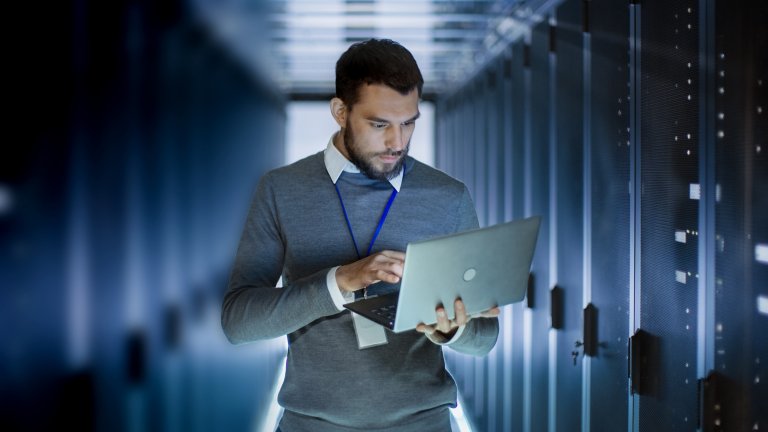 A Life Sciences professional reviews data on his laptop while standing near a row of servers.