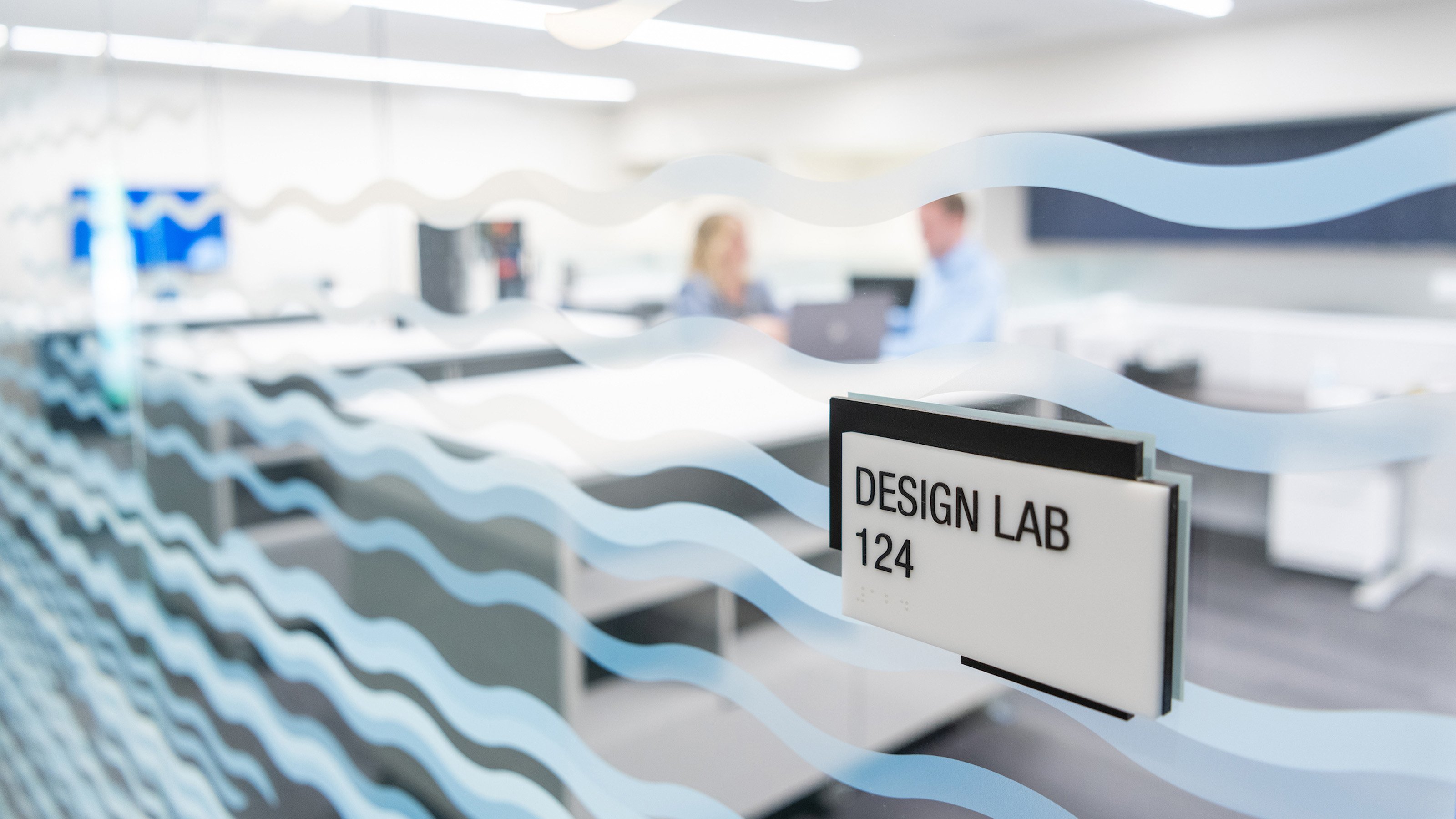 A male and female electrical safety engineer are shown behind a frosted window in a design lab as they discuss safety circuit engineering plans for their industrial manufacturing facility.