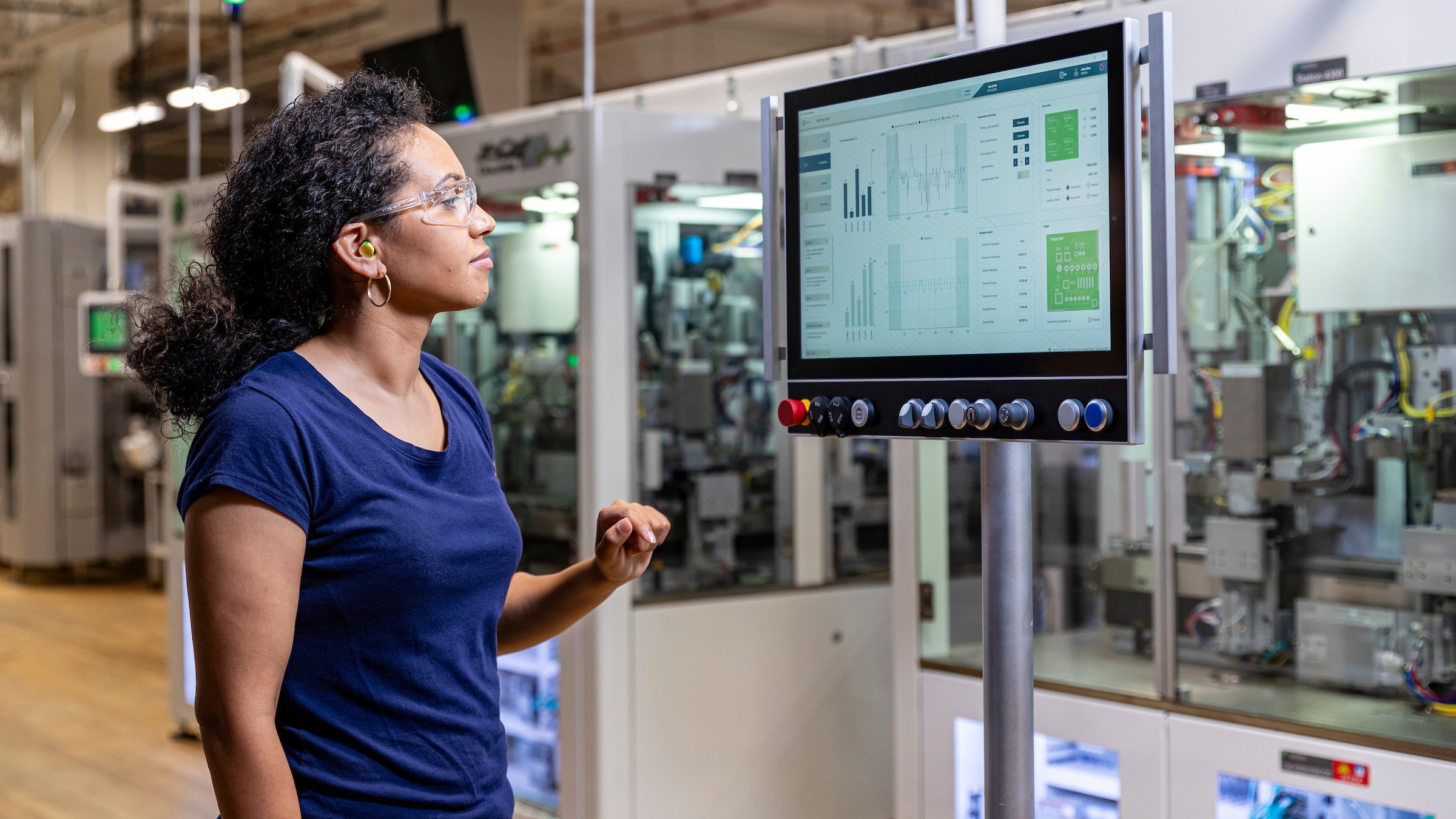 Eine Bedienerin mit Schutzbrille blickt auf eine OptixPanel-Anzeige vor einer Montageanlage.