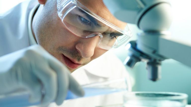 A lab worker at a life sciences facility pours solution into a petri dish positioned under a microscope.